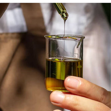 Joy Garden Jasmine Hair Serum liquid texture: a golden botanical oil elixir being poured into a beaker, showing the weightless and high-shine formula.
